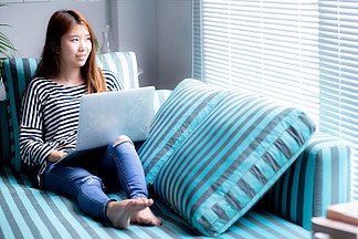 美麗的肖像年輕亞洲女性在客廳沙發上使用筆記本電腦休閑，女孩在網上與筆記本自由職業者一起工作，擁有快樂、溝通的商業理念。