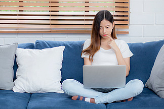 美麗的年輕亞洲女性在筆記本電腦上工作，在家沙發上思考想法，自由職業女孩坐在沙發上使用筆記本在客廳上網，一個人，生活方式概念。