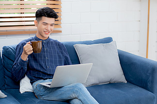 年輕的亞洲商人微笑著在家工作，在客廳的沙發上使用筆記本電腦在線工作，自由職業者在沙發上使用筆記本和喝咖啡，新常態，生活方式概念。