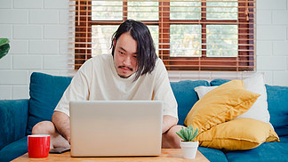 在家工作的亞洲自由職業者，在客廳沙發上用筆記本電腦創作的男性。商業青年業主企業家，玩電腦，在現代住宅概念下檢查工作場所的社交媒體。