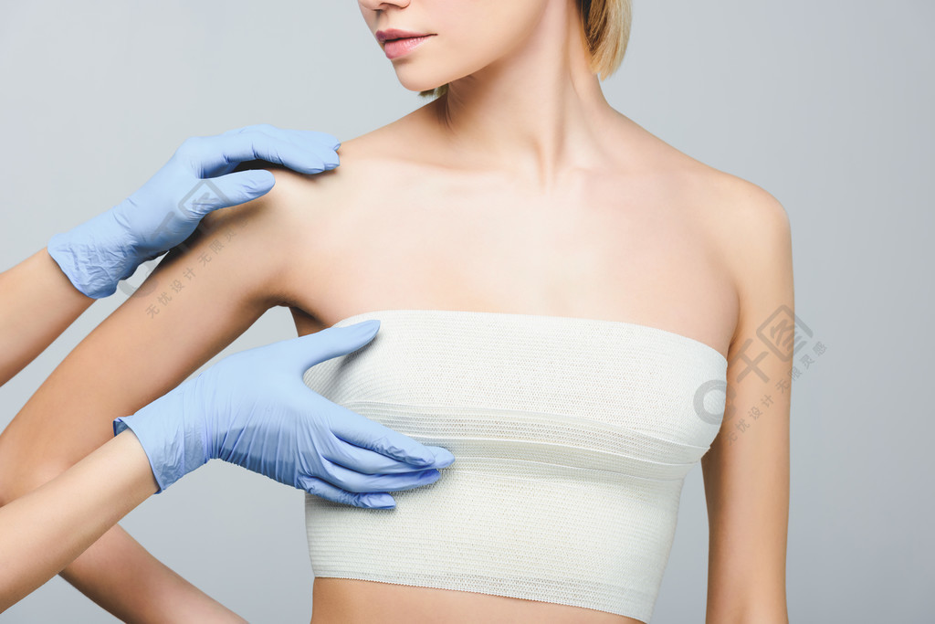 裁剪视图整形外科医生在乳胶手套检查女性乳房绷带 隔离在灰色