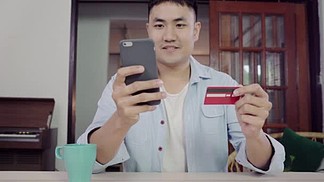 亚洲男<i>子</i>使用智能手机在线购物和信用卡在网上的客厅家庭。亚洲人坐在餐桌旁休闲服