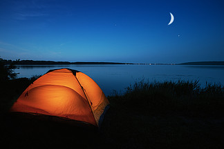 湖边点亮的橙色旅游帐篷。夜空。隐私、旅行与和谐的概念。湖边点<i>燃</i>的橙色旅游帐篷