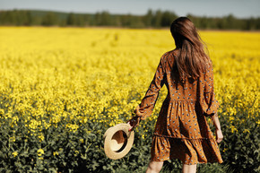 时尚的年轻女子在黄色的花朵领域。戴着草帽，穿着花裙子的女孩。题字的地方。后视图。背景与黄色的花朵和蓝天。