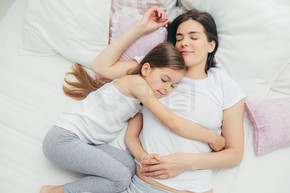 家庭和睡觉时间的概念。高兴的昏昏欲睡的母亲和她可爱的女儿在床上拥抱，做着美梦，躺在白色的床上，有良好的关系，互相照顾