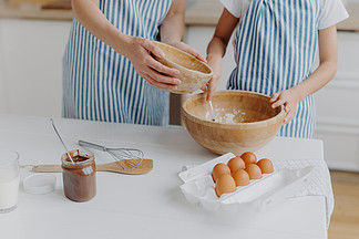 母亲和女儿用手混合配料准备面团和烘<i>烤</i>美味糕点的裁剪图像，站在厨房桌旁，放着鸡蛋，在玻璃中融化巧克力，穿着条纹蓝色围裙