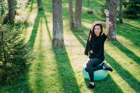 活跃灵活的黑发女人的照片用健身球伸展，做普拉提或健身运动，在阳光明媚的春日在森林里的绿草上摆姿势，在新鲜空气中享受体操运动。