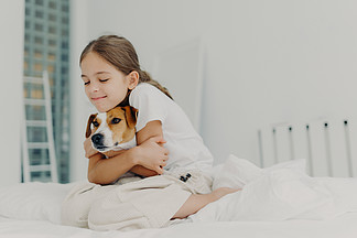高兴的小女孩和宠物玩耍，拥抱狗，闭上眼睛，穿着睡衣，醒来后在床上摆姿势，表达对纯种动物的爱。床上用品概念