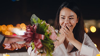 年轻的亚洲夫妇惊喜时刻与玫瑰花束在城市之夜的屋顶餐厅浪漫晚餐与<i>甜</i><i>蜜</i>的时刻庆祝周年。爱情关系，结婚的概念。