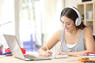 一名学生在线学习的肖像，他戴着耳机和笔记本电脑，在家里办公桌前的笔记本上记笔记