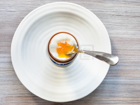 在灰色木板上的白盘上用勺子在杯子里煮软煮的棕色鸡蛋的顶部视图