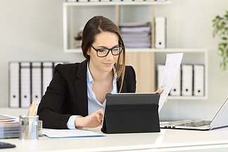 戴眼镜的办公室工作人员将报告与办公桌上的平板电脑内容进行比较