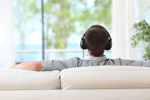 一个男人放松和听音乐的背影,他戴着耳机坐在沙发上,透过家里的窗户看