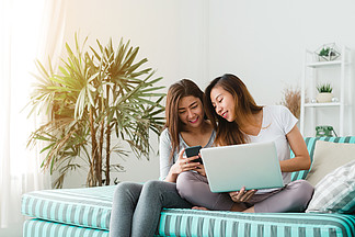 美丽的年轻亚洲女性 LGBT 女同性恋幸福夫妇坐在沙发上，在家客厅使用笔记本电脑、电脑和电话在线购买。 Lgbt 女同性恋夫妇一起在室内概念。