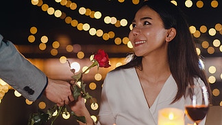 年轻的亚洲夫妇在城市夜晚的屋顶餐厅浪漫晚餐时用玫瑰花惊喜时刻，<i>甜</i><i>蜜</i>的时刻庆祝周年。爱情关系，结婚的概念。