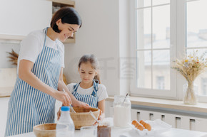 一张快乐的妈妈和孩子在厨房一起做饭的照片，穿着围裙准备好吃的东西，做食物，用打蛋器打鸡蛋，在家烤糕点。牛奶，鸡蛋，巧克力，桌上的面粉