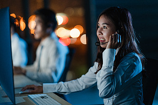 Millennial Asia 年轻的呼叫中心团队或客户支持服务主管在深夜办公室使用计算机和麦克风耳机工作技术支持。电话营销或销售工作概念。