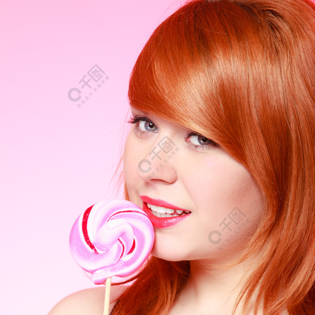 性感的年轻女子拿着糖果redhair女孩在粉红色上吃甜棒棒糖工作室拍摄