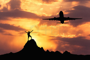 飞机和一个站立的快乐人的剪影。日落时，飞机和站在山峰上的快乐男人和举起双臂的剪影。夏季景观，降落客机在橙色天空中飞翔，云朵和登山者