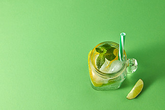 绿色背景上带有酒<i>精</i>鸡尾酒的玻璃罐的顶部视图。柠檬和酸橙、冰、水和玻璃中的塑料吸管的柑橘类水果片。冷酒<i>精</i>或非酒<i>精</i>夏季饮料的概念。用绿色背景上的塑料吸管天然柑橘类水果自制的闪闪发光的冷酒<i>精</i>莫吉托。顶视图。