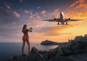 美丽的女人在日落时拍摄着陆飞机的照片。夏天的风景，女孩站在岩石上，在五颜六色的天空下飞行客机。女人和降落的商用飞机。