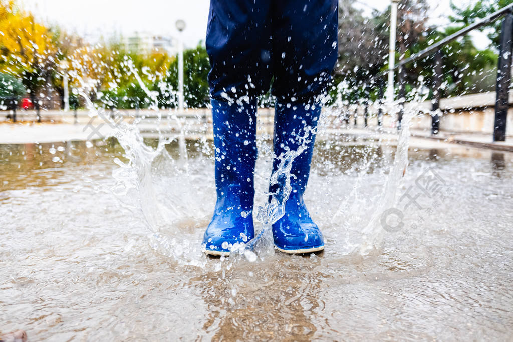 孩子穿着蓝色雨靴在公园里的水坑里溅水joaquincorbalancom