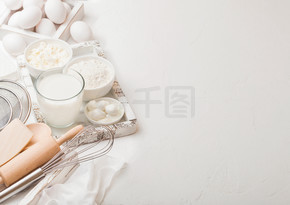 白色桌子背景上的新鲜乳制品。一杯牛奶、一碗面粉、干酪和鸡蛋。一盒烘焙用具。在老式木盒中搅拌和抹刀。顶视图。