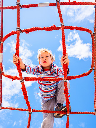 金发男孩栖息在儿童游乐场的网状绳梯结<i>构</i>上，享受攀爬乐趣。 joaquincorbalan.com