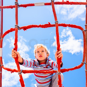 金发男孩栖息在儿童游乐场的网状绳梯结构上，享受攀爬乐趣。 joaquincorbalan.com