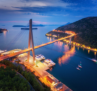 美丽的现代桥梁在晚上的鸟瞰图。克罗地亚杜布罗夫尼克。日落时的道路、<i>船</i><i>只</i>、游艇和城市灯光的顶视图。与港口、山、高速公路、蓝色海和多云天空的夏天风景