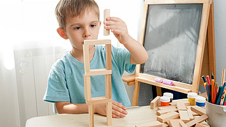 快乐微笑的男孩在幼儿园教室的桌子上用木块和砖块建造塔。锁定和自我隔离期间聪<i>明</i>儿童和家庭教育的概念。快乐微笑的男孩在幼儿园教室的桌子上用木块和砖块建造塔。锁定和自我隔离期间聪<i>明</i>儿童和家庭教育的概念。