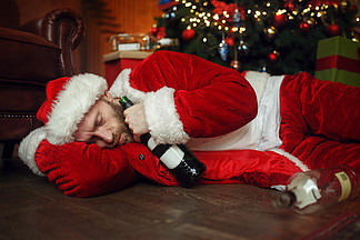 醉酒的圣诞老人带着一瓶酒睡在圣诞树下，讨厌的派对，幽默。不健康的生活方式，穿着节日服装的大胡子男人，新年和酗酒。醉酒的圣诞老人睡在圣诞树下