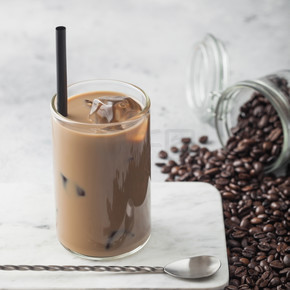 一杯冰咖啡加牛奶在大理石板上，一罐咖啡豆和长勺放在浅桌背景上。