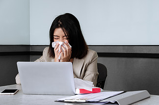 生病的女商人拿着笔记本电脑坐在办公桌前，在会议室工作时用面部纸巾打<i>喷</i>嚏和咳嗽。冠状病毒covid-19在工作场所的传播和风险