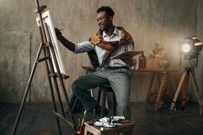 微笑的画家用画笔和调色板坐在画架上。男艺术家在他的工作场所画画，在车间创作大师作品。在画架上用画笔和调色板微笑的画家