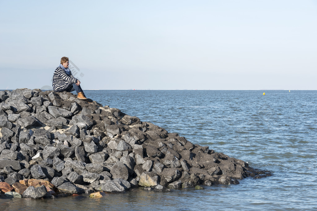 穿着棕色靴子和蓝色衣服的成年白人女性坐在海边的岩石上望向地平线