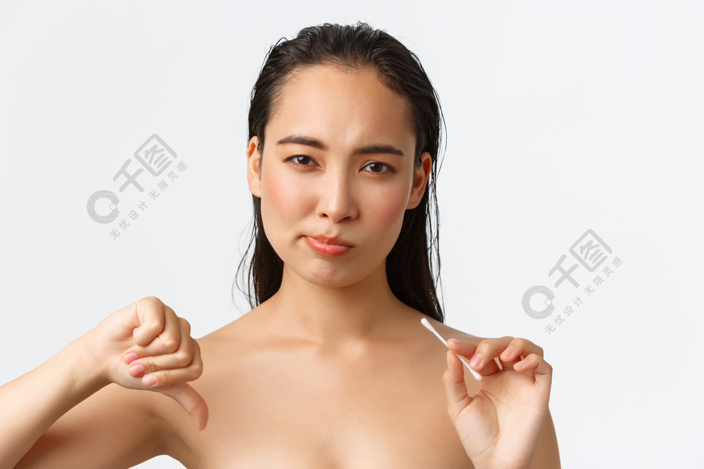 护肤、女性美容、卫生和个人护理概念。不满美丽的亚洲女人赤身裸体地站着棉花芽，做鬼脸，做出不喜欢、不赞成的大拇指手势。护肤、女性美容、卫生和个人护理概念。不高兴的美丽亚洲女人赤身裸体地站着，拿着棉花芽，
