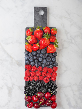 新鲜的有机夏季浆果混合在大理石背景的黑色大理石板上。覆盆子、草莓、蓝莓、黑莓和樱桃。顶视图