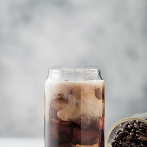 一杯黑冰咖啡，配上新鲜牛奶和一罐生咖啡豆和咸焦糖，放在浅桌背景上。