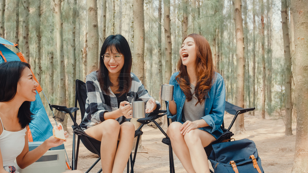 一群年轻的亚洲露营者朋友坐在森林帐篷旁的椅子上。少女旅行者在夏日的露营地放松和交谈。户外活动、冒险旅行或假期。