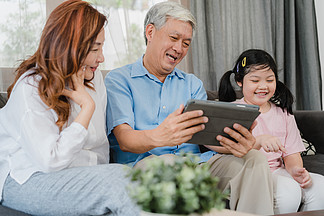 亚洲祖父母和孙女在家里使用平板电脑。中国老人、爷爷奶奶快乐地度过家庭时光，与小女孩一起查看社交媒体，躺在客厅的沙发上
