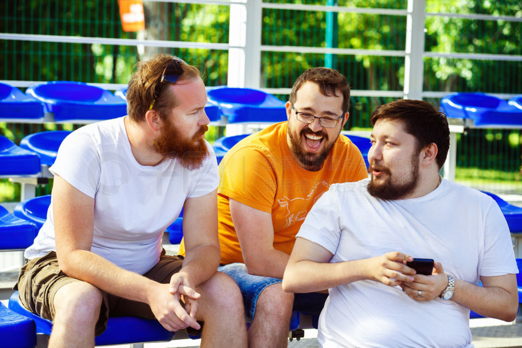 Three male friends are watching mobile phone video and laughing sitting at summer day at stadium bac