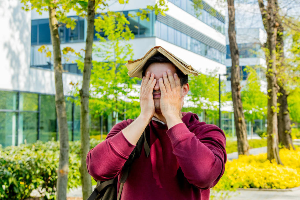 Young upset student with book on head stay near modern building