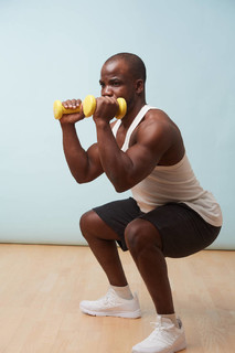 英俊的黑色大胆男子穿无袖白色衬衫做蹲与两个浅黄色拼贴塑料哑铃。 淡蓝色背景。 健身锻炼。