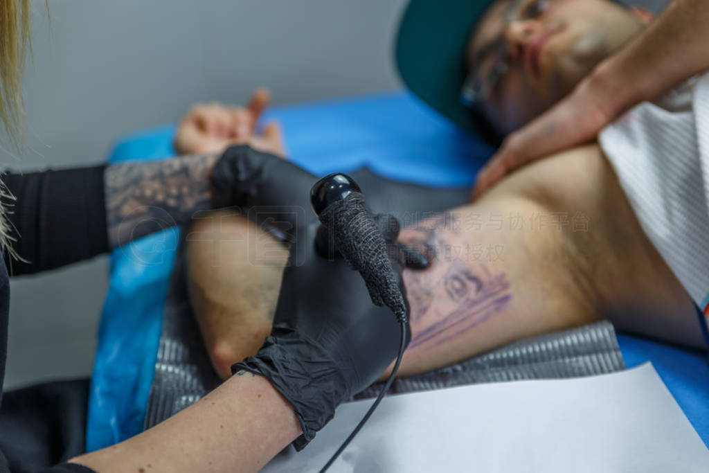 A boy with glasses and blue cap is tattooed by a young woman in