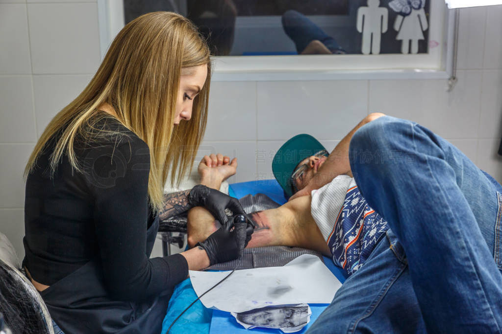 A boy with glasses and blue cap is tattooed by a young woman in