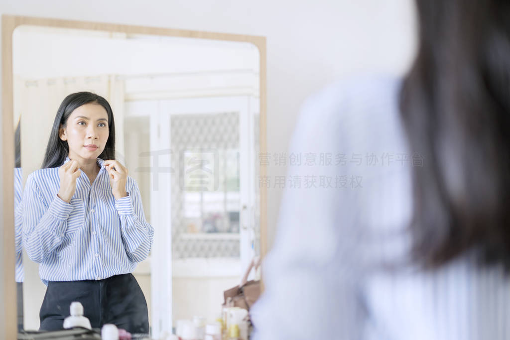 Elegant Asian businesswoman buttoning up shirt in front of mirro