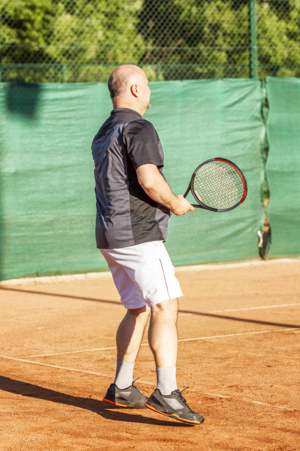 Adult bald man plays tennis on the court. Sunny day.