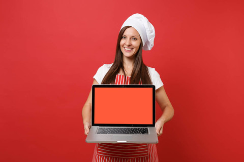 家庭主妇女厨师或面包师穿条纹围裙，白色t恤，厨师帽子隔离在红墙背景上。女士手提电脑空白屏幕的宣传内容模拟复制空间概念