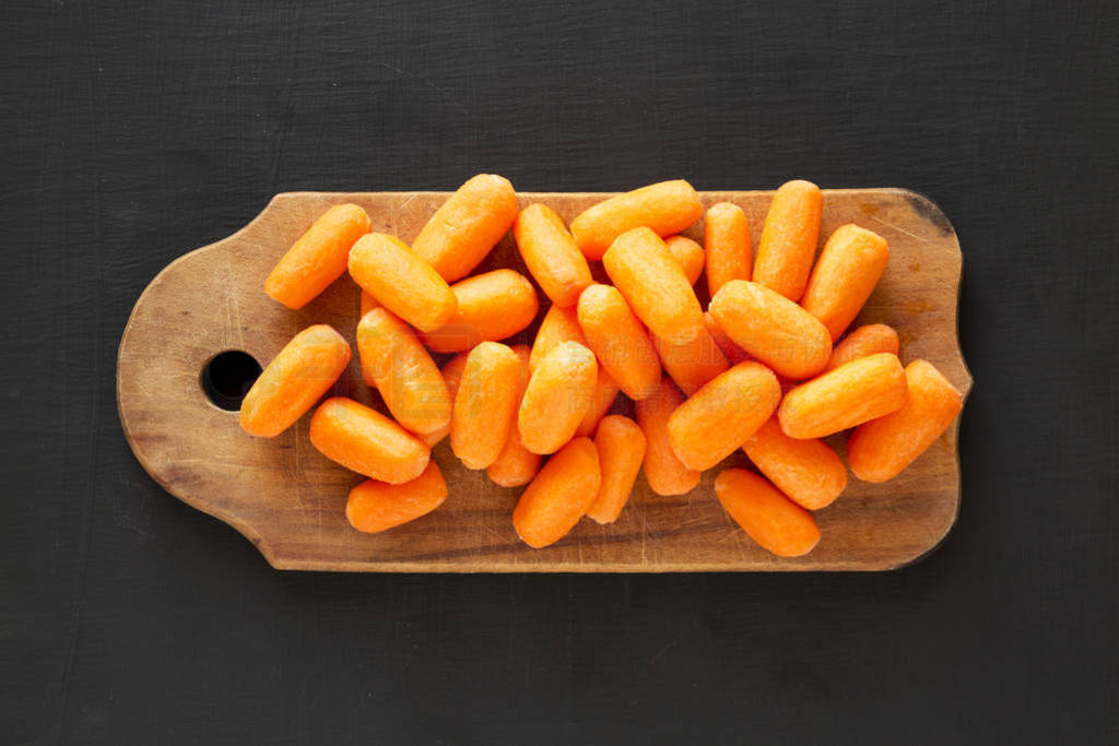 Fresh baby carrots on rustic wooden board on black surface, top